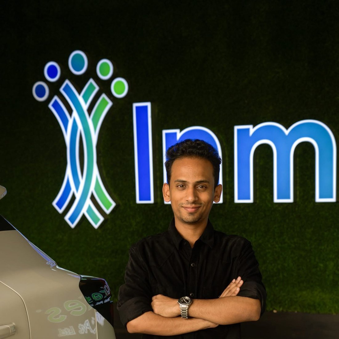 Ajmal Basheer smiling with arms crossed in front of the illuminated Inmakes logo on a green wall