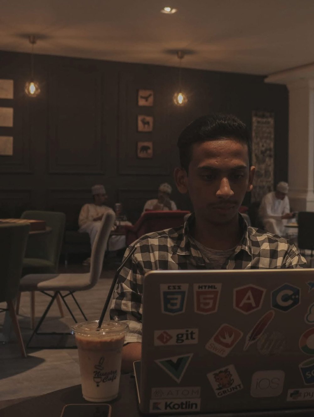 Ajmal Basheer coding on a sticker-covered laptop in a café, iced coffee beside him