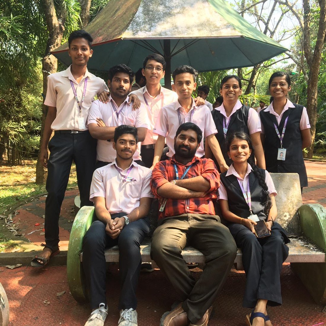 Ajmal Basheer with his college team in uniform and lanyards, gathered around a bench under a garden umbrella on campus