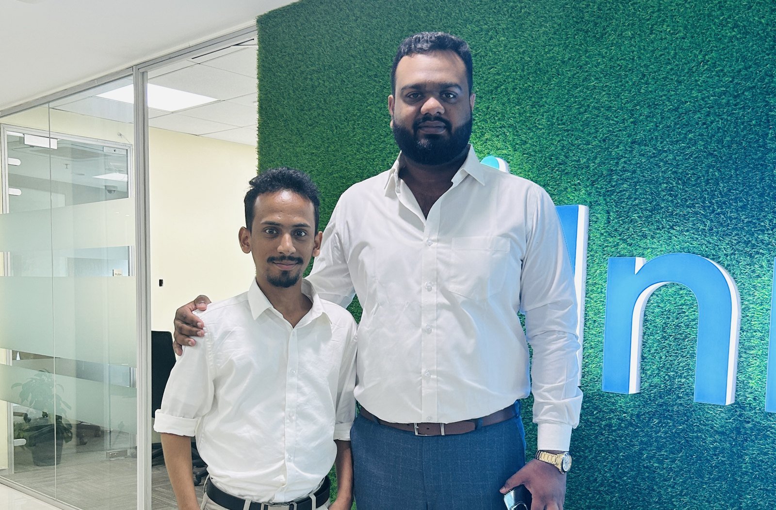 Ajmal Basheer standing with JM Bilal, Founder and CEO of JBIS Group of Companies, his arm around Ajmal's shoulder, in front of a green office wall