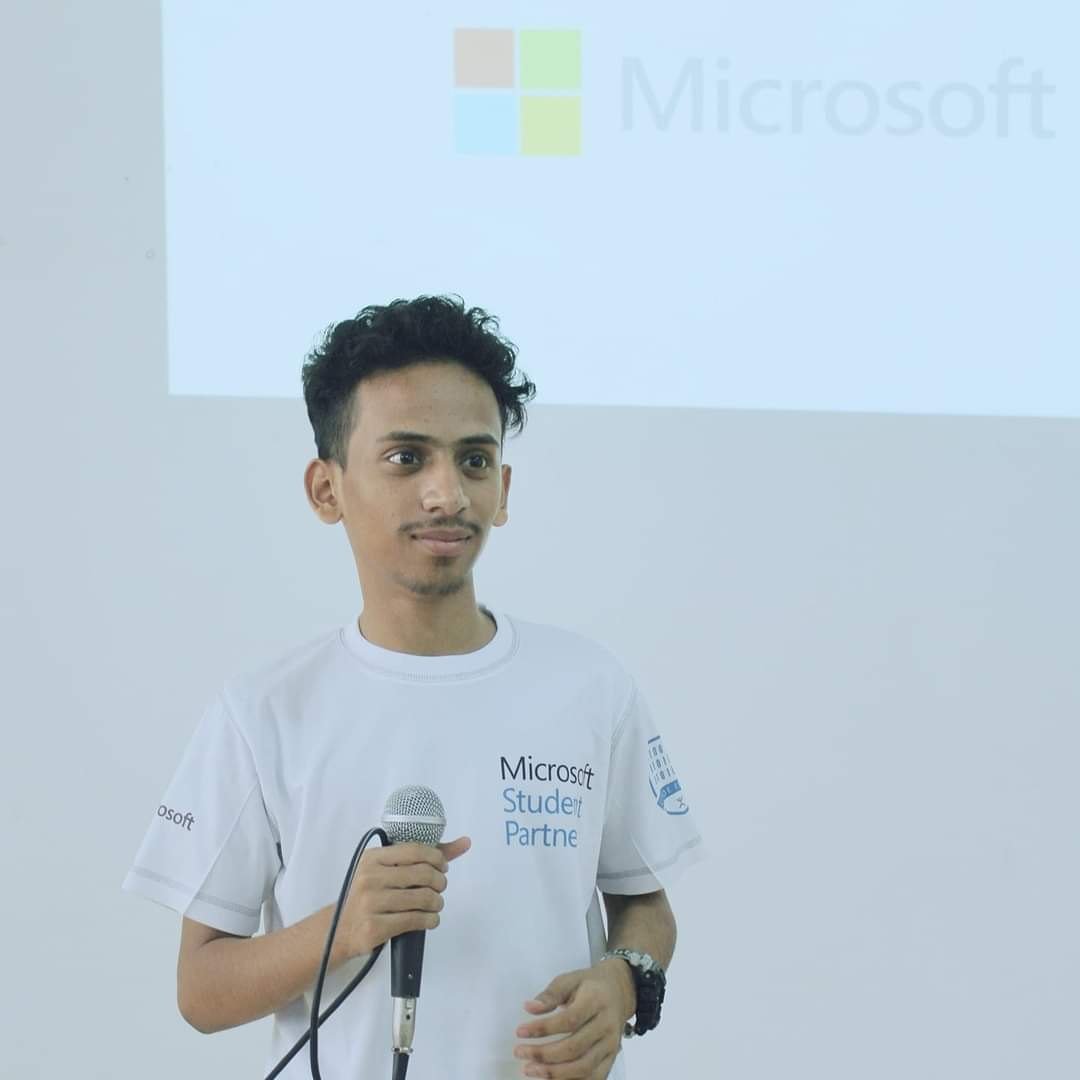 Ajmal Basheer in a Microsoft Student Partner T-shirt, holding a microphone in front of a projected Microsoft logo